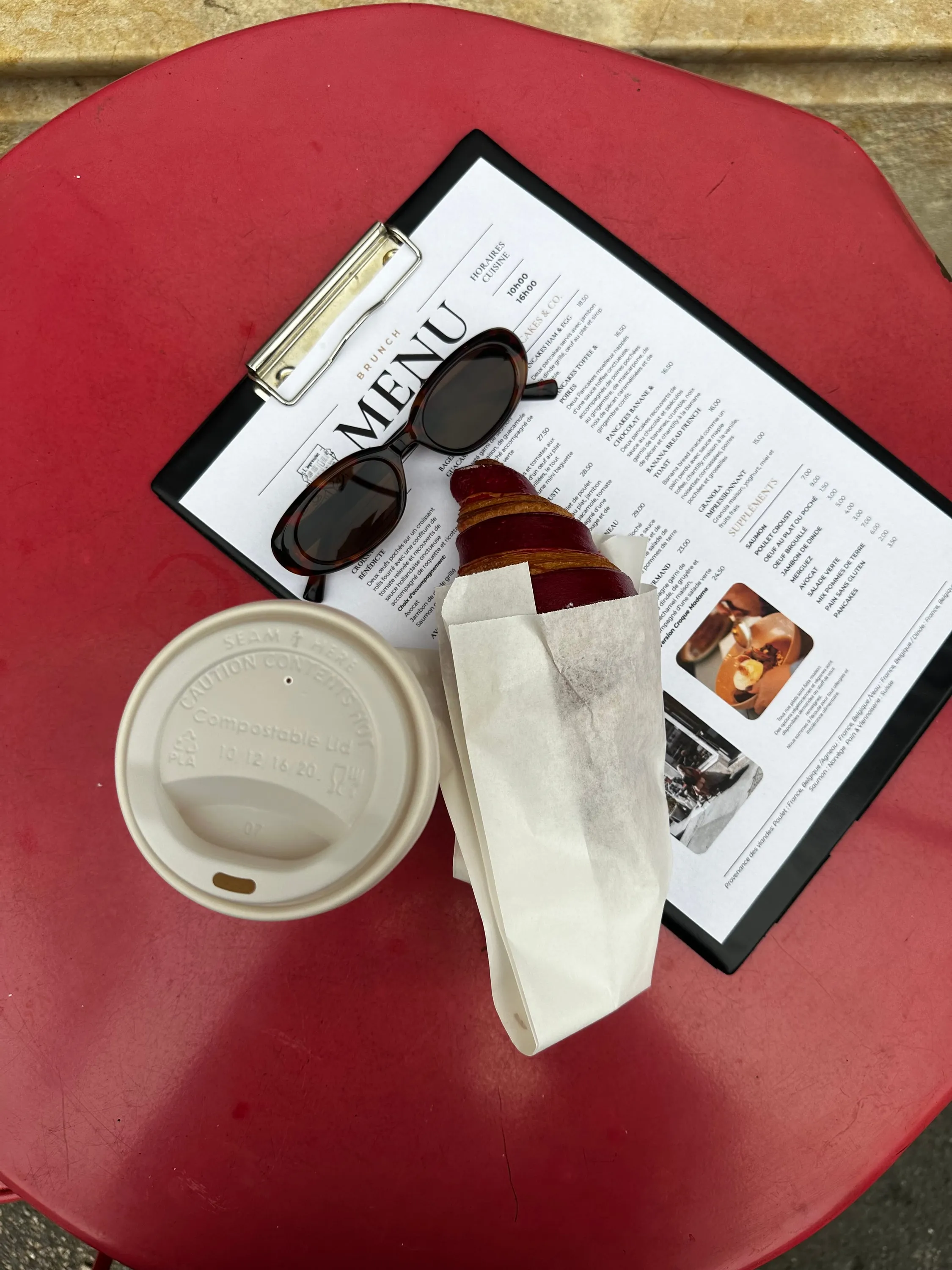 The brunch menu on its clipboard, croissant and takeaway coffee, editorial flat lay.
